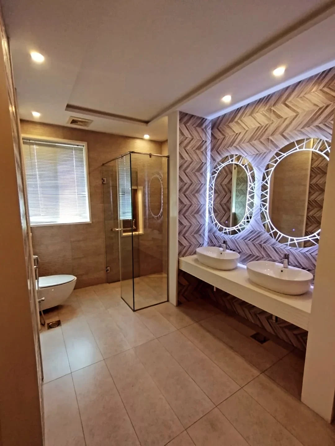 Luxury master bathroom with dual vessel sinks, LED oval mirrors, herringbone wall tiles and glass shower cabin in Lake City Lahore house