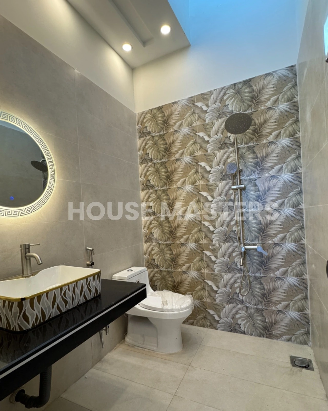Premium Bathroom Design – 5 Marla House, Bahria Orchard Stylish bathroom with leaf-pattern accent wall, black countertop, and illuminated round mirror in 5 Marla luxury home, Bahria Orchard.